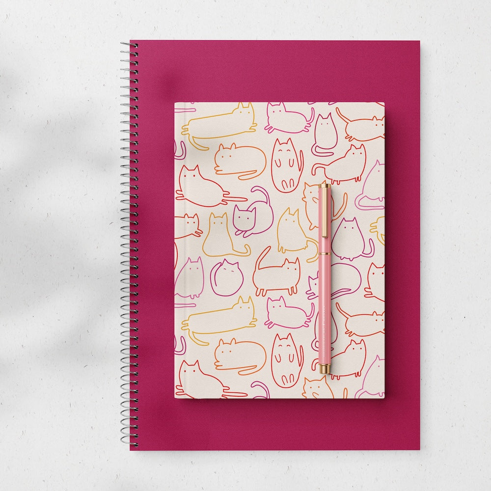 Hardcover notebook with cartoon cats in pink, yellow and orange on a cream background. On top of a maroon spiral notebook with a pink pen